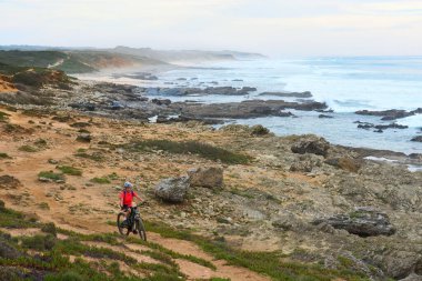 nice senior woman riding her electric mountain bike at the rocky and sandy coastline of the atlantic ocean in Porto Covo, Portugal, Europe