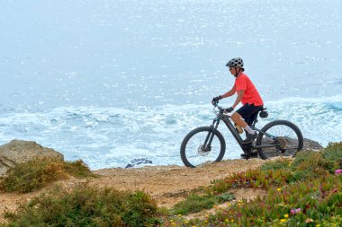 nice senior woman riding her electric mountain bike at the rocky and sandy coastline of the atlantic ocean in Porto Covo, Portugal, Europe