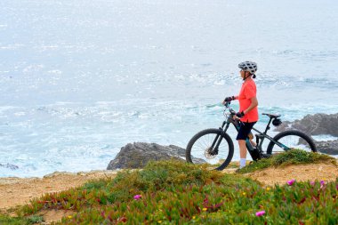 nice senior woman riding her electric mountain bike at the rocky and sandy coastline of the atlantic ocean in Porto Covo, Portugal, Europe