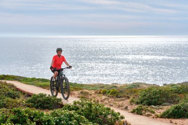 nice senior woman riding her electric mountain bike at the rocky and sandy coastline of the atlantic ocean in Porto Covo, Portugal, Europe