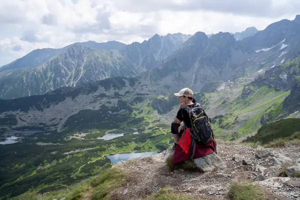 Bir kadın dağın tepesinden aşağı bakıyor, yürüyüş kıyafetleri giymiş, beline kırmızı bir yağmurluk bağlanmış, sırtında bir sırt çantası.