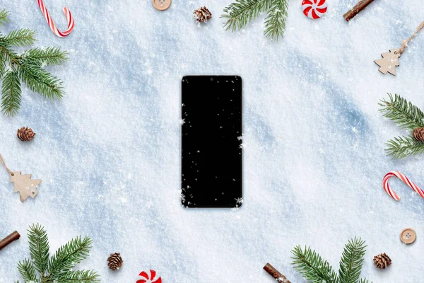 Smart phone with blank screen in snow surrounded by Christmas decorations