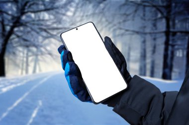 Mobile phone in hand with winter glove. Trees covered with snow in background. Isolated display for app promotion
