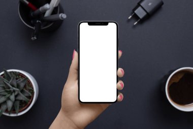 Model için izole edilmiş görüntüsü olan kadın eli telefon. İş, uygulama ya da web sayfası sunumu için ofis modeli. Arka planda kahve fincanı, bitki, kalem, şarj aleti olan ofis masası