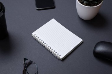 Siyah çalışma masasındaki boş not defteri kopyalama alanı. Ofis malzemeleri, telefon, bardak, kahve fincanı, bitki. Düşük hafif iş bileşimi