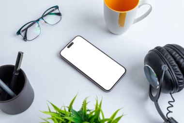 Ofis masasına, kahve fincanı, bardak, kalem, bitki ve kulaklıkla çevrili uygulama sunumu için izole edilmiş ekranlı telefon modeli.