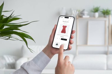 Akıllı telefonuyla internetten alışveriş yapan bir kadın. Ecommerce web sayfasından kırmızı akış elbisesi almak. Oturma odası arka planda.