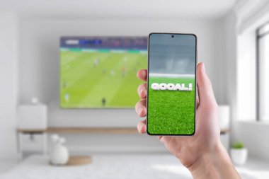 Telefon ekranındaki hedef. Spor sonuçlarını internette takip etme kavramı. Oturma odasında arka planda televizyon ve futbol maçı var.