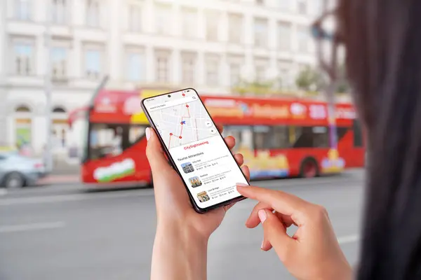 City Sightseeing app on smartphone in woman hands. City tourist bus in background