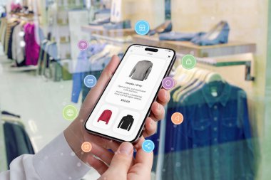 Kadının elleri, alışveriş simgeleriyle çevrili bir akıllı telefonu tutarken, bir butikte arka planda giysiler sergileniyor.