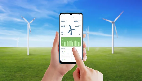 Hand utilizes a wind generator app to monitor energy production. Meadow with spinning wind generators in background