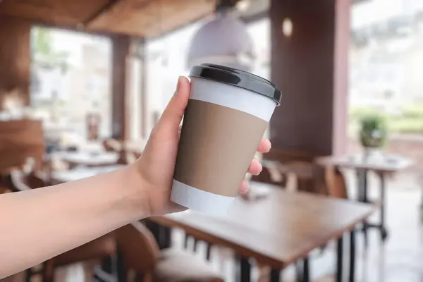 Coffee paper cup held in hand with coffee shop in the background. Space for logo promotion