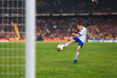 Genç futbolcu topa kararlılıkla vuruyor, dolu bir stadyumda heyecanlı bir maçın parlak ışıkları altında gol atıyor.