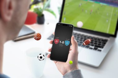 Eldeki cep telefonu bir uygulamada eşleşen sonuçları gösteriyor, arka planda dizüstü bilgisayarda bir futbol maçı gösteriliyor. Çeşitli spor topları telefonun etrafında uçuyor, dinamik spor bağlantılarını vurguluyorlar.