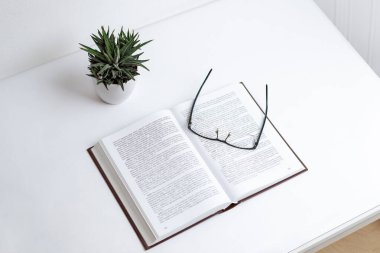 Yoğun metin ile açık kitap üzerinde dinlenen bardaklar, öğrenmeyi, okumayı ve bilgi edinmeyi sembolize eder; bir bitki odaklı minimalist masa ayarı ve sakinliği ekler