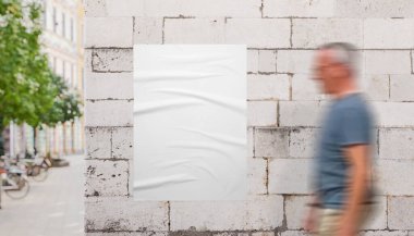 Şehir kordonunun yakınındaki parlak taş duvarda beyaz poster modelinin yanından geçen bir adam. Poster, marka ve kampanya modelleri için ideal.