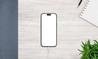 Şarjlı akıllı telefonun en üst görüntüsü boş ekranlı, defter ve bitkiyle birlikte masanın üzerine yerleştirilmiş. Üretkenlik ve uygulama modellemeleri için ideal