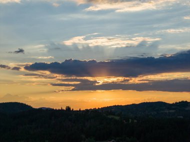 Sıcak günbatımı ışığı katmanlı bulutları kırıp uzak ormanlık tepeler ve vadiler boyunca altın renkler bırakıyor.
