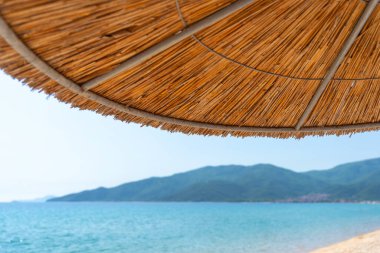 Saman plajı şemsiyesi, güneşli bir yaz gününde mavi denizin ve uzak dağların manzarasını gölgeliyor.