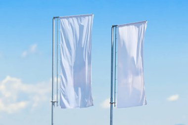Açık mavi gökyüzüne karşı metal direkler üzerinde sallanan iki uzun dikey beyaz bayrak, minimal açık hava reklam kavramı