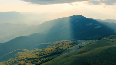 Alp vadileri ve yüksek tepeler üzerinden bulutları aşan dramatik güneş ışınları yakalayan 4K hava videosu.