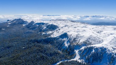 Jahorina dağının görkemli hava manzarası parlak mavi gökyüzünün altında taze karla kaplı, ormanlık yamaçlar ve panoramik alp manzarası sergiliyor.