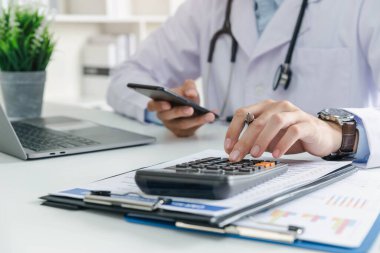 Doktor, klinikteki hesap makinesini kullanarak sağlık giderlerini ve gelirlerini hesapladı..