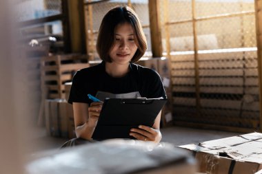 Kadın inşaat deposu işçisi malları kontrol ediyor. Panoda eşyaların bir listesi var ve kadın onları işaretliyor gibi görünüyor.