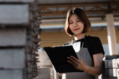 Kadın inşaat deposu işçisi malları kontrol ediyor. Panoda eşyaların bir listesi var ve kadın onları işaretliyor gibi görünüyor.