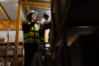 Bir depoda iki işçi, biri sarı yelek giyiyor. Kadının elinde bir tablet, adamın elinde de bir dosya var.