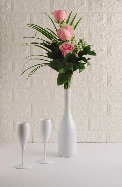 Still life composition made of white champagne bottle, glasses and bouquet of pink rose flowers on the table. White party background. Valentine's Day concept.
