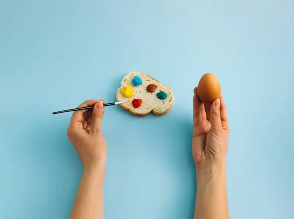 An boiled egg in hand and slice of bread with colorful palette on pastel blue background. Woman is coloring eggs for Easter holiday. Creative Easter composition. Flat lay, top view.
