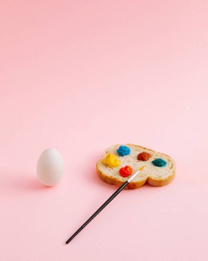 Beyaz yumurtadan yapılan soyut Paskalya kompozisyonu ve renkli paletli bir dilim ekmek. Pastel pembe arka plan. Üst manzara, düz yatış. Boşluğu kopyala.