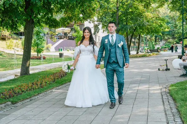 Güzel bir gelin ve damat açık havada huzurlu bir anın tadını çıkarıyor. Genç çift el ele yürüyorlar, arka planda doğa ile çevrili romantik bir yürüyüşü paylaşıyorlar..
