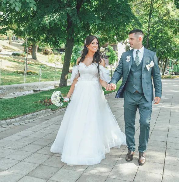 Güzel bir gelin ve damat açık havada huzurlu bir anın tadını çıkarıyor. Genç çift el ele yürüyorlar, arka planda doğa ile çevrili romantik bir yürüyüşü paylaşıyorlar..