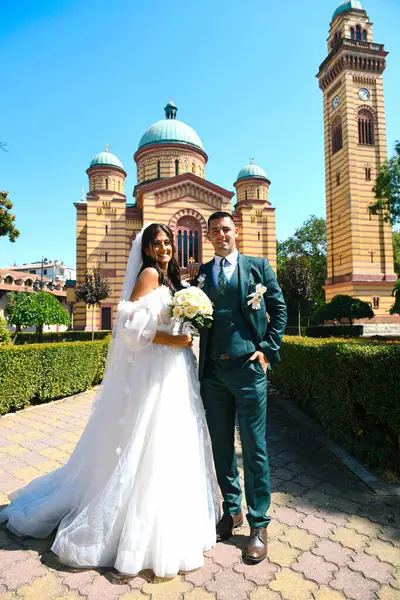 Kilisenin önünde duran evli bir çift. Geleneksel kilise düğünü. Gelin çok güzel bir elbise giymiş, elinde bir buket gül çiçeği tutuyor..