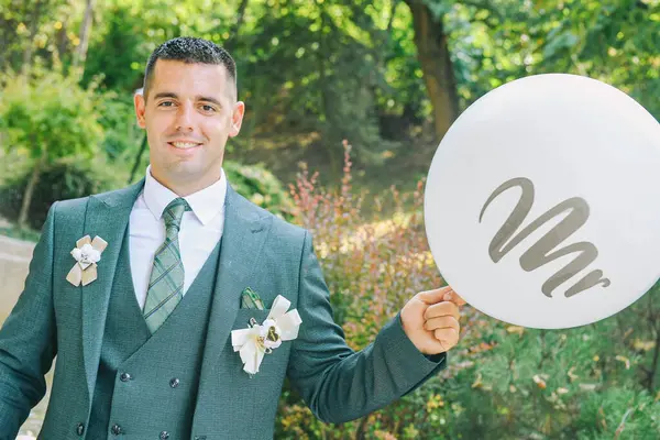 Bir damat parkta duruyor, zarif yeşil bir takım elbise giyiyor ve elinde büyük bir balon tutuyor. Gülümsüyor ve anın tadını çıkarıyor.. 