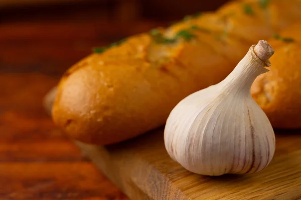 Close-up of a head of garlic with garlic bread with aromatic herbs on a wooden board in the background.