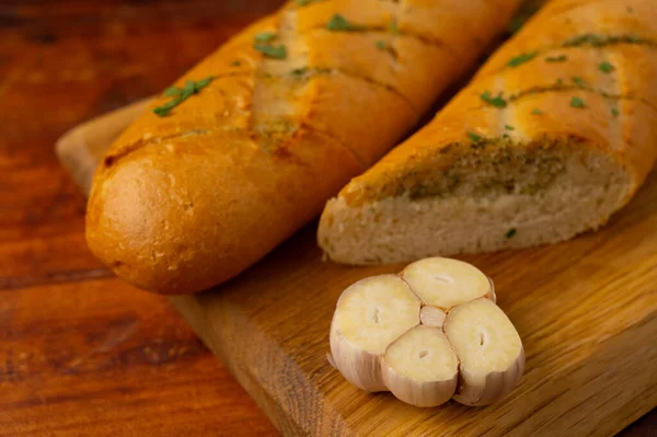 Close-up of fresh cheese garlic bread cut in half with aromatic herbs on a wooden board  on a wooden table with sliced garlic in front.