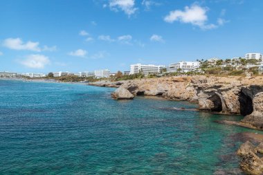 Ayia Napa tatil beldesi. Tatilciler için bir cennet. Mavi gökyüzü ve beyaz bulutlarla güneşli bir gün. Otellerin panoramik manzarası, mavi göl ve kaya ve mağaralı kıyı şeridi. Kıbrıs. Akdeniz.