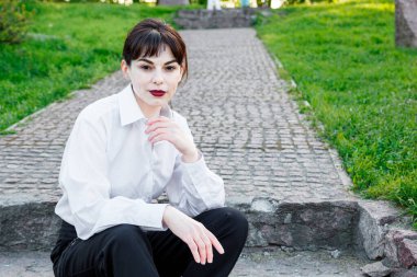 Beyaz tişörtlü, siyah pantolonlu, arka planında pantolon askısı olan yeşil çimenler olan güzel bir kız.