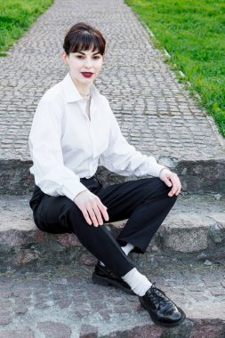 Beyaz tişörtlü, siyah pantolonlu, arka planında pantolon askısı olan yeşil çimenler olan güzel bir kız.