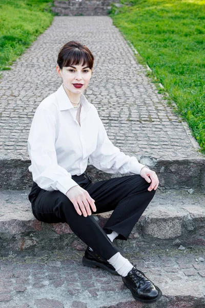 A beautiful girl in a white shirt, in black trousers with suspenders against the background of the sky and green grass close-up