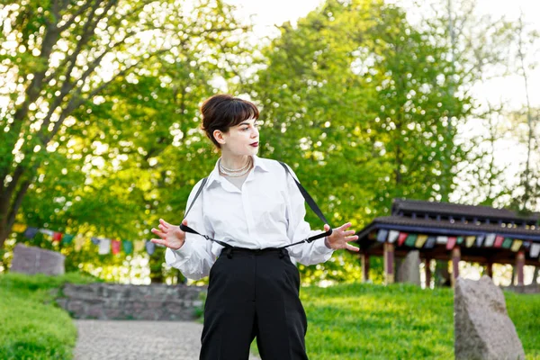 A beautiful girl in a white shirt, in black trousers with suspenders against the background of the sky and green grass close-up