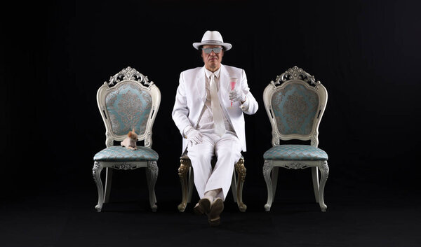  богатый джентльмен в белом костюме сидит на стуле, черный студийный фон