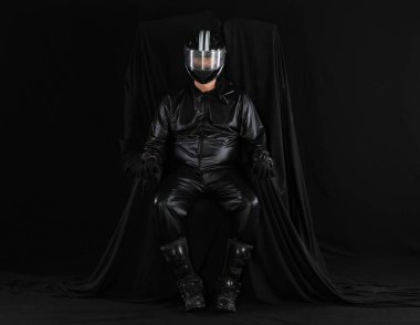 Siyah arka planda bir sandalyede kasklı bir motorcunun portresi.