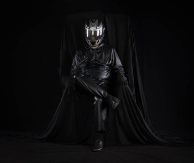 Siyah arka planda bir sandalyede kasklı bir motorcunun portresi.