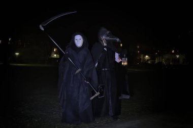 Two figures dressed as death and plague doctor at night in the park under a lantern