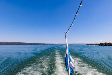 View from a ship over the white and blue Bavarian flag over Ammersee lake in Bavaria with sailboats on the water and Ammergau Alps in the background