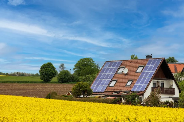 stock image Single-family house with photovoltaic modules on the roof in front of a rape field in full bloom under a blue sky